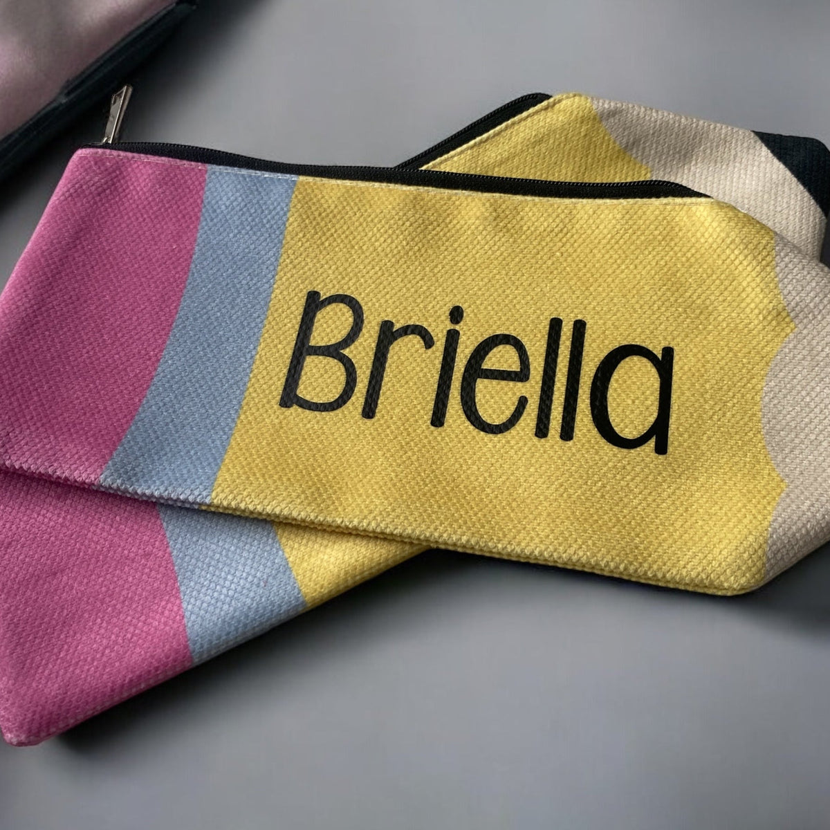 A beige canvas pencil pouch with the name 'Briella' printed in yellow, laying on a desk with a collection of colorful pencils.