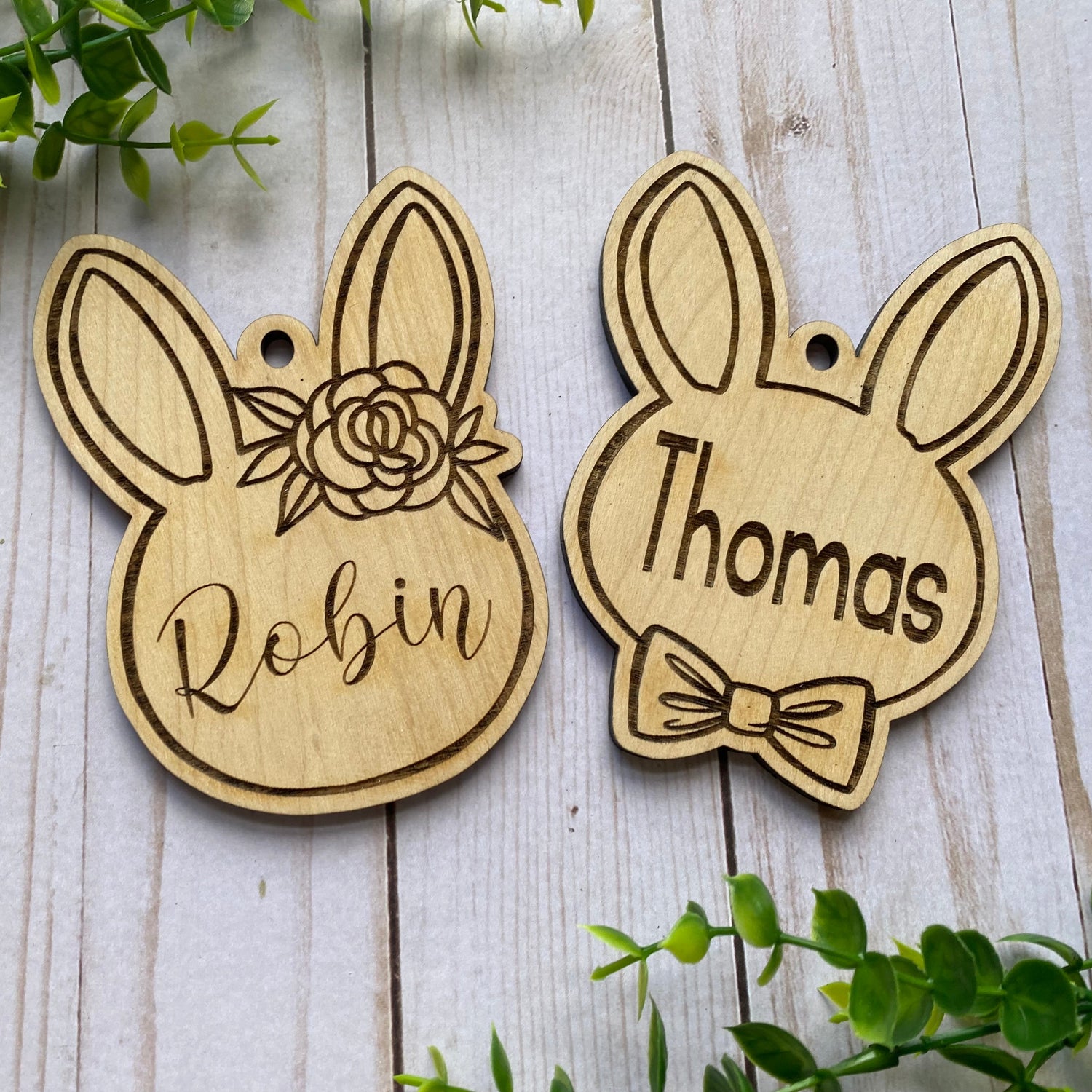 Two wooden bunny-shaped tags with engraved names on a wooden surface with greenery.